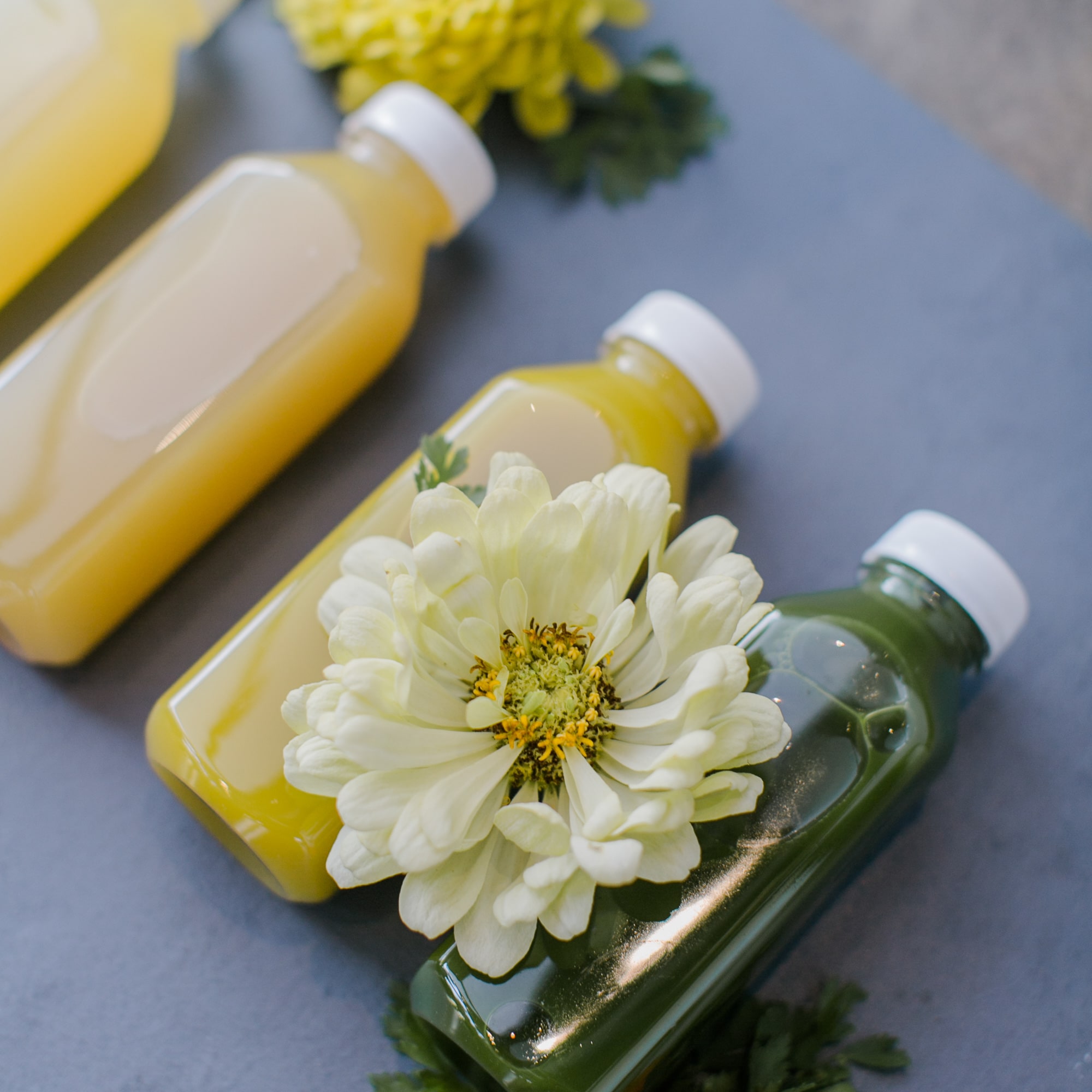 Close-up of glass juice bottles in yellow and green hues topped with a white flower on a gray surface, fresh and clean in mood