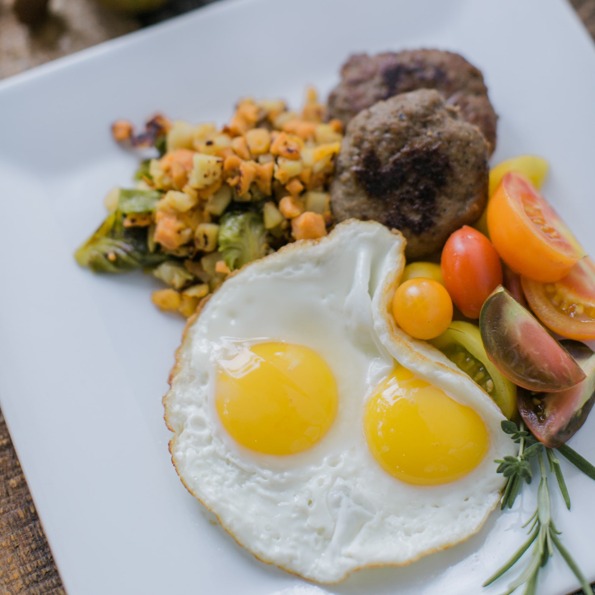 Overhead breakfast plate with two sunny-side-up eggs, sausage patties, diced potatoes, and cherry tomatoes