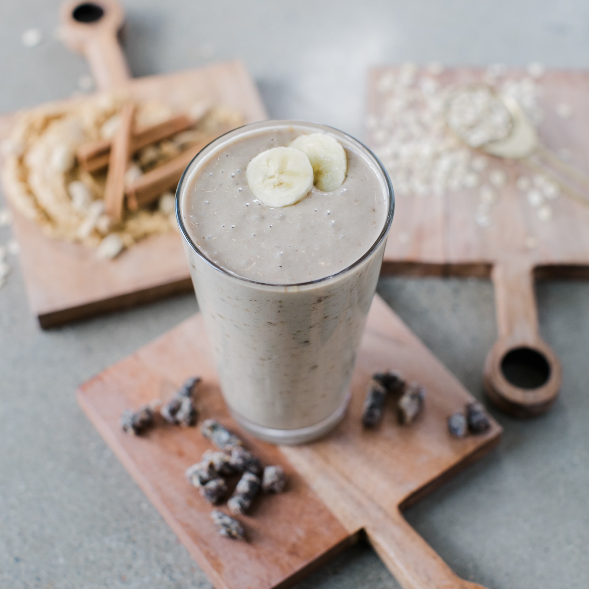 A tall glass of beige smoothie topped with banana slices, set on wooden boards with cinnamon sticks, oats, and small bowls, soft natural light, fresh and wholesome mood.