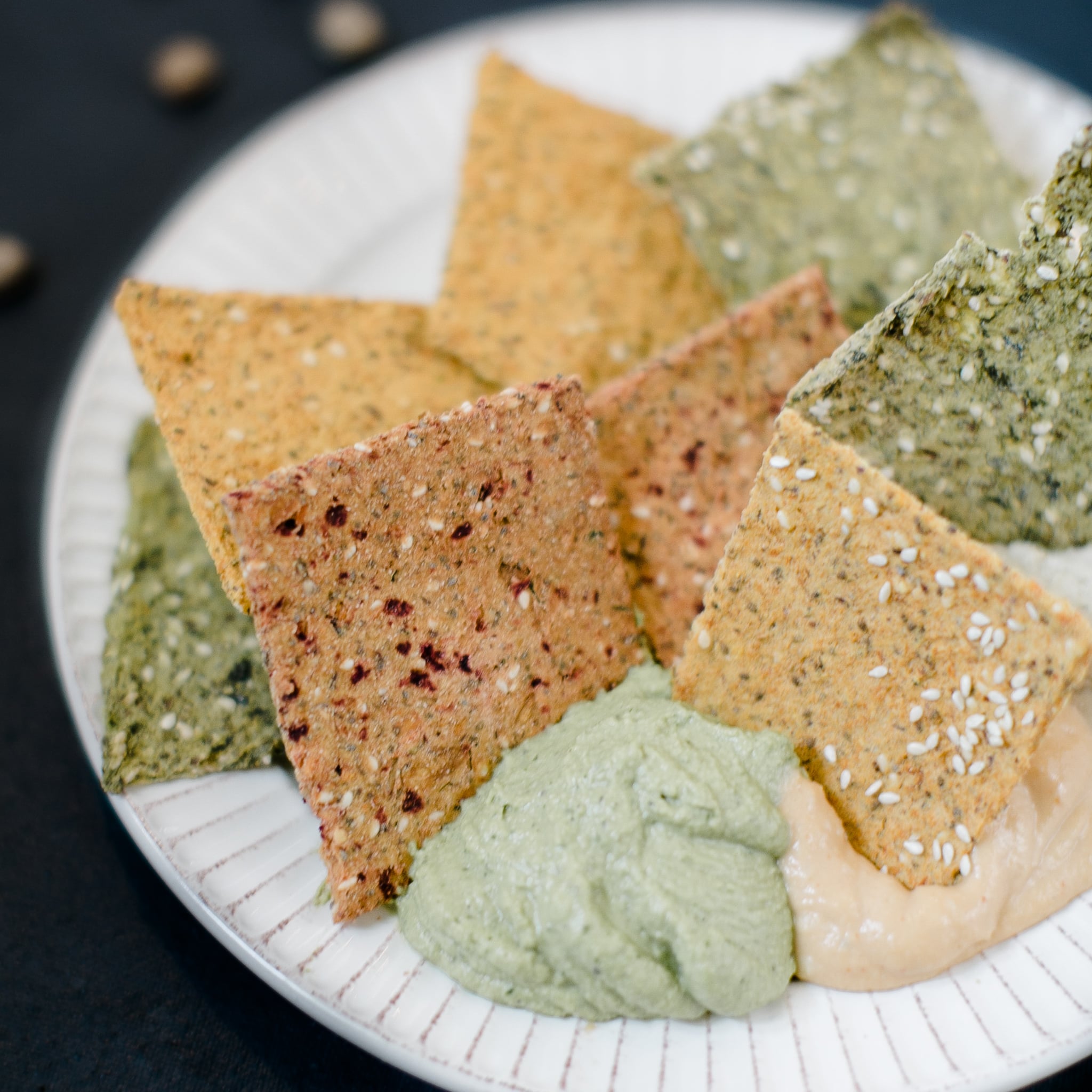 Overhead close-up of assorted seeded crackers in orange, green, and red-speckled varieties arranged on a white plate, warm and natural light, rustic and wholesome mood.