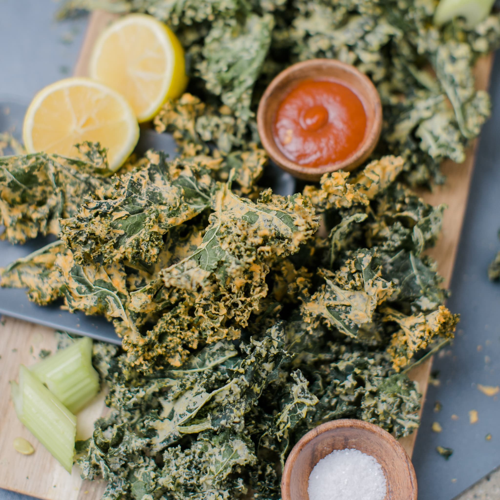 Close-up of crispy seasoned kale chips with orange seasoning, lemon halves, celery sticks, and a small bowl of dip, bright and zesty mood with fresh greens.
