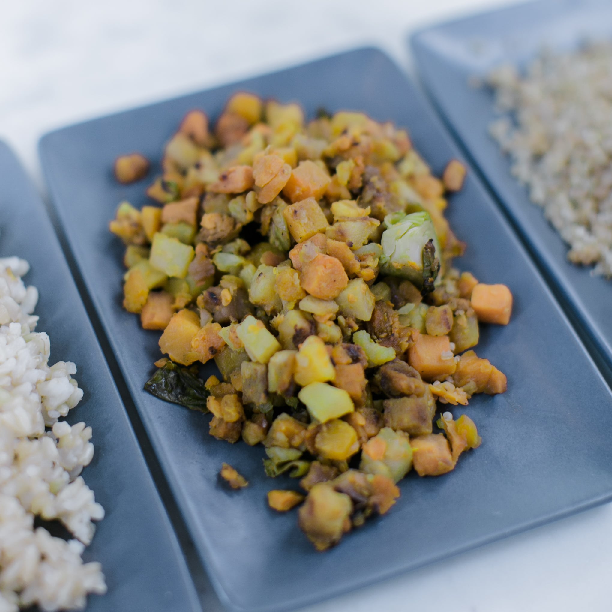 Close-up of diced roasted vegetables including sweet potato and brussels sprouts on a dark plate with rice visible, warm earthy tones, comforting and nourishing mood.