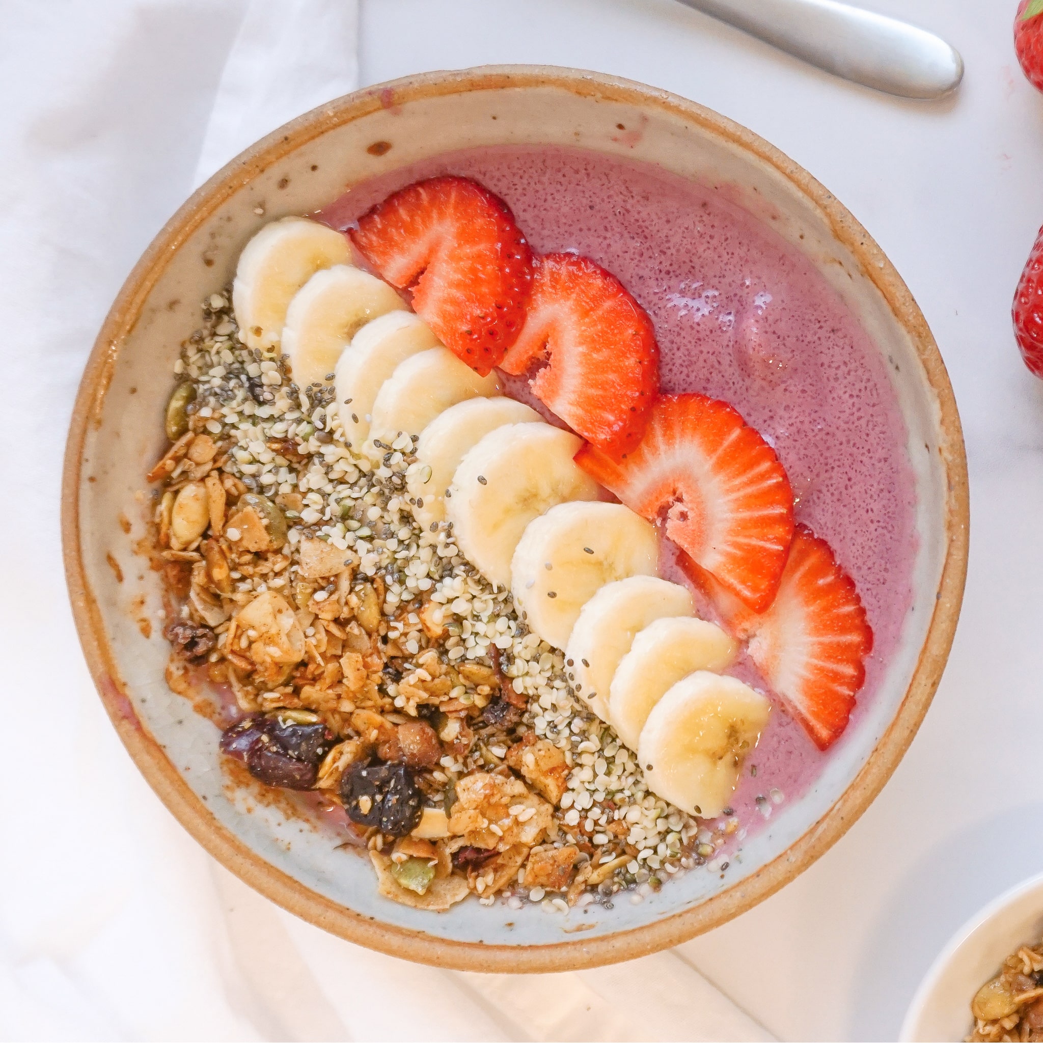 Overhead shot of a bowl of pink smoothie topped with banana slices, strawberries, granola and seeds, bright and fresh