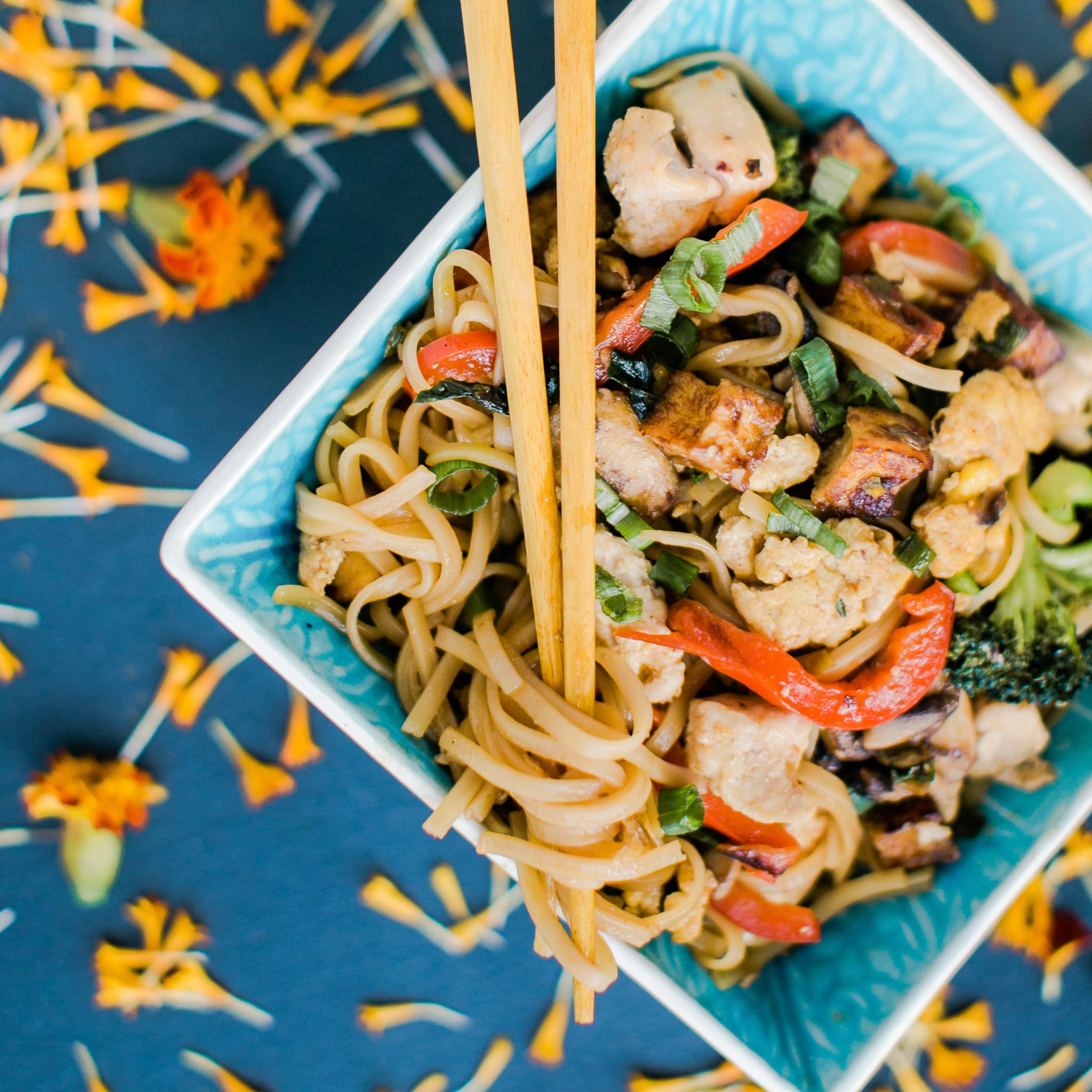 Overhead close-up of a noodle stir-fry with chicken, peppers, and scallions in a blue bowl, chopsticks resting on top, warm kitchen lighting, appetizing and home-style mood.